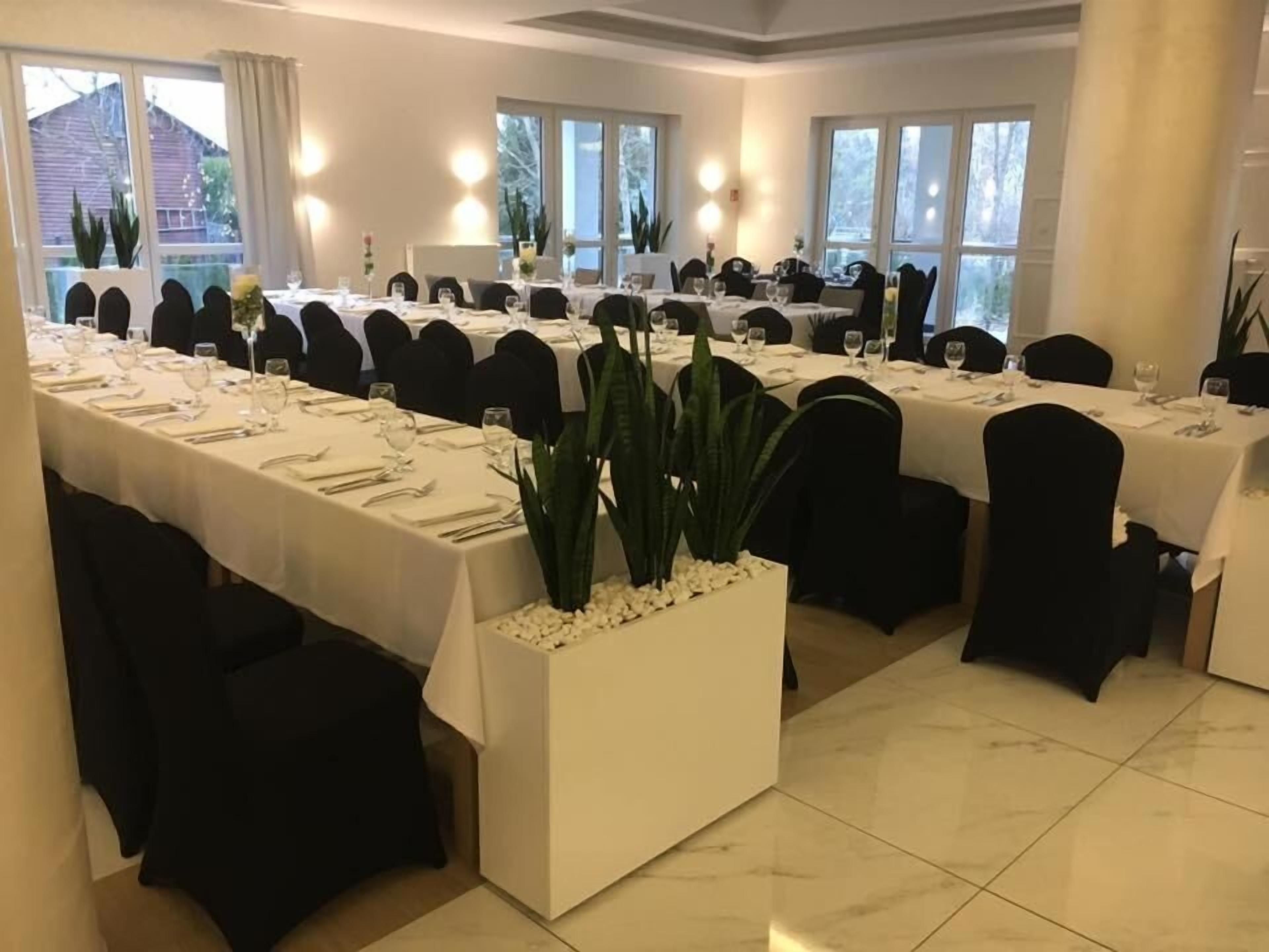 Un comedor formal con varias mesas dispuestas para una comida, ventanas amplias y plantas en macetas.