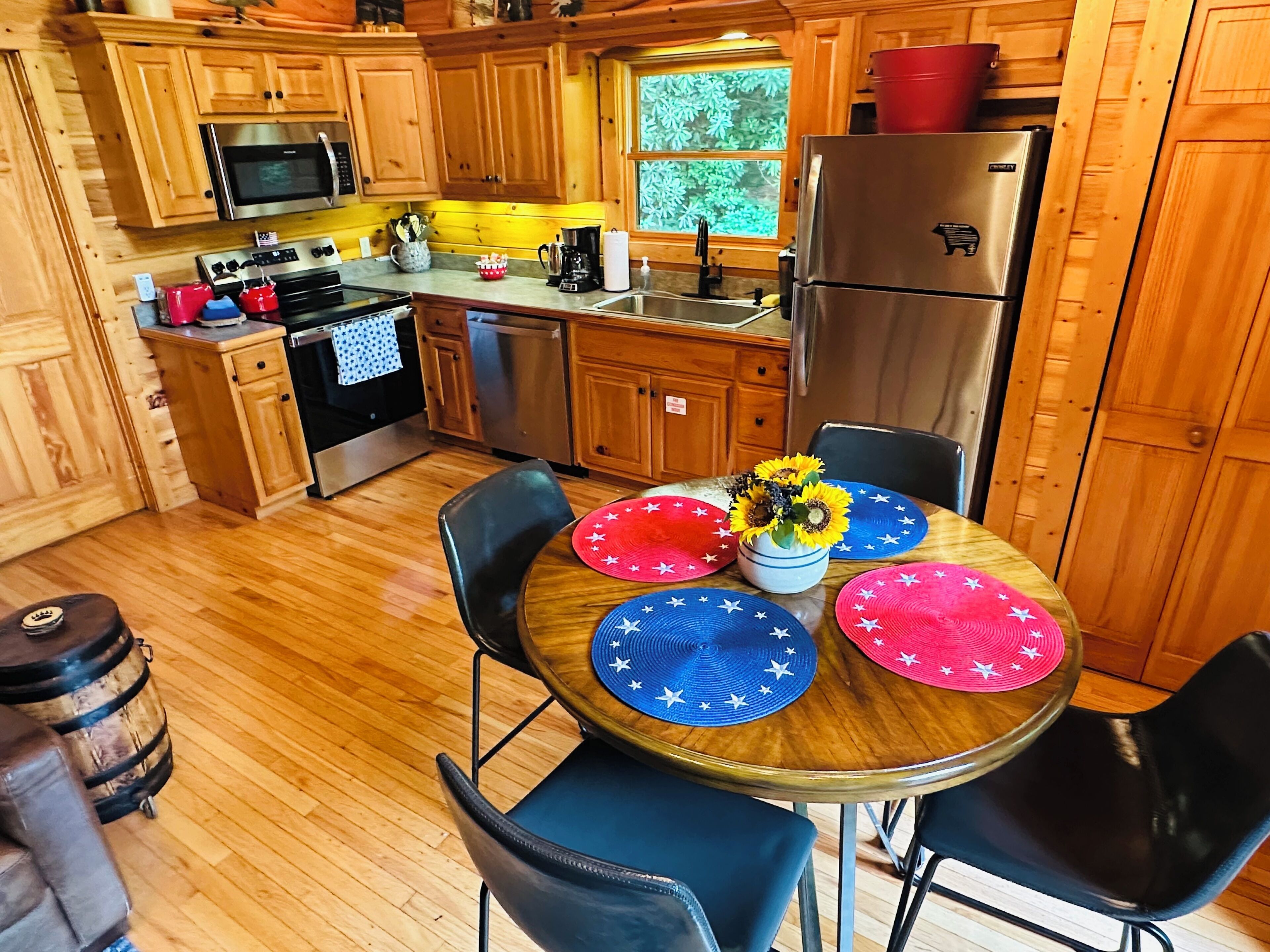 A wooden kitchen with a dining table set for four, a refrigerator, and a window with a view of greenery.