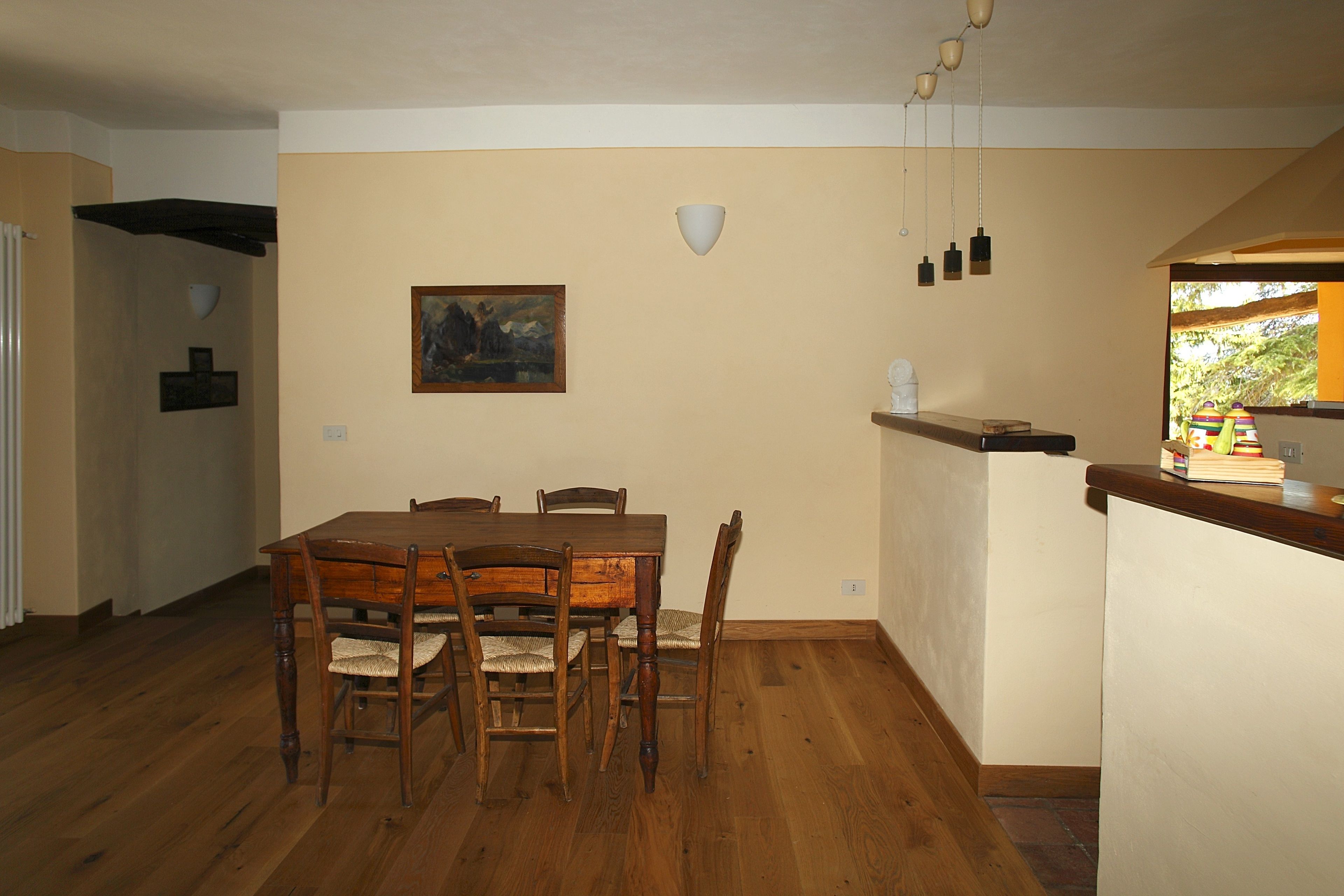 A dining area with a wooden table and chairs, a painting on the wall, and a kitchen counter with a window.