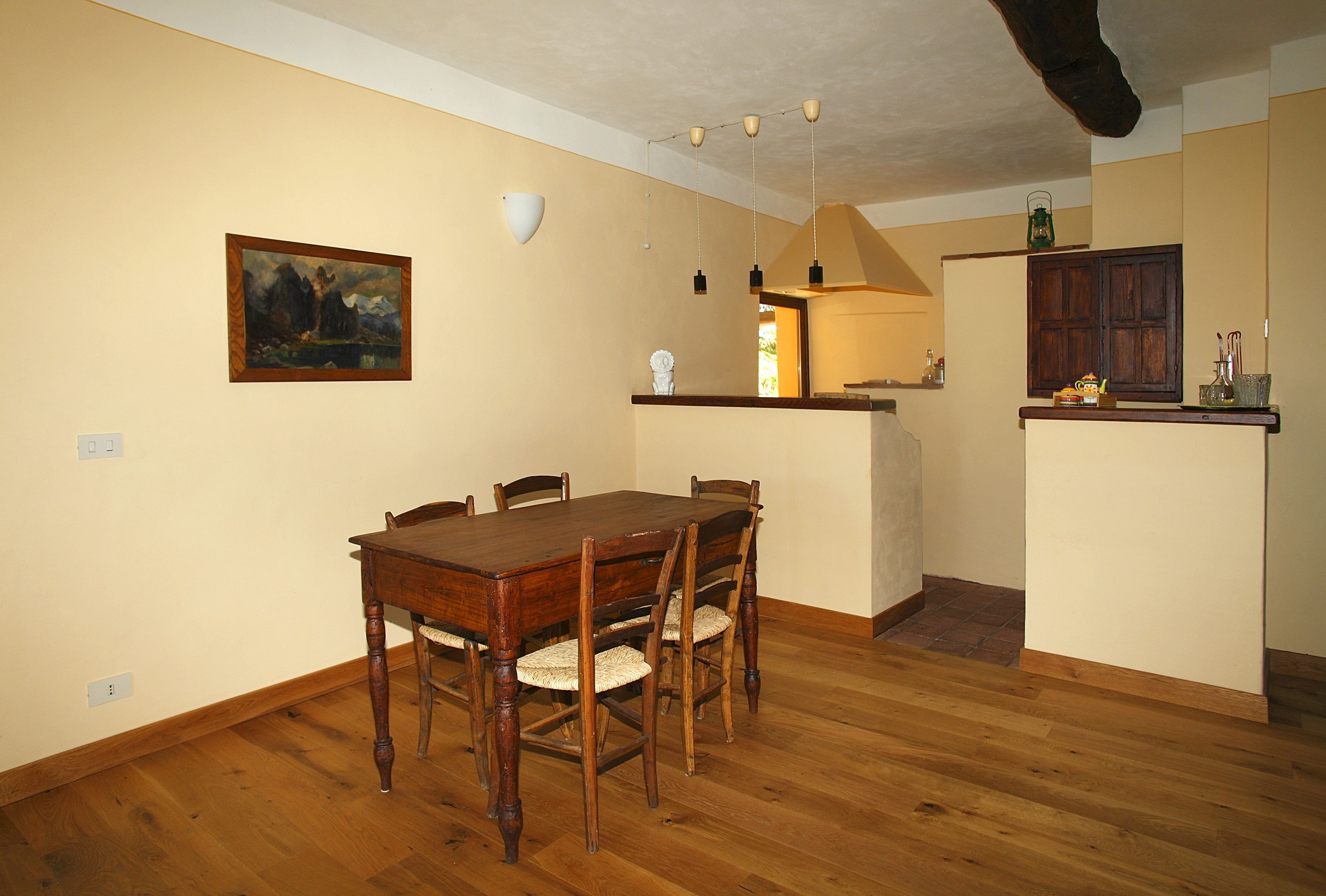 Uma área de refeições com mesa e cadeiras de madeira, um quadro na parede e uma cozinha com armários e bancada.