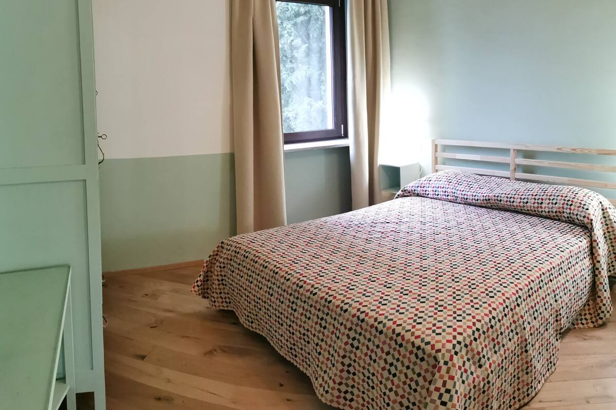 Un dormitorio con suelo de madera, una cama con colcha estampada, una ventana con cortinas color beige y una pared verde.