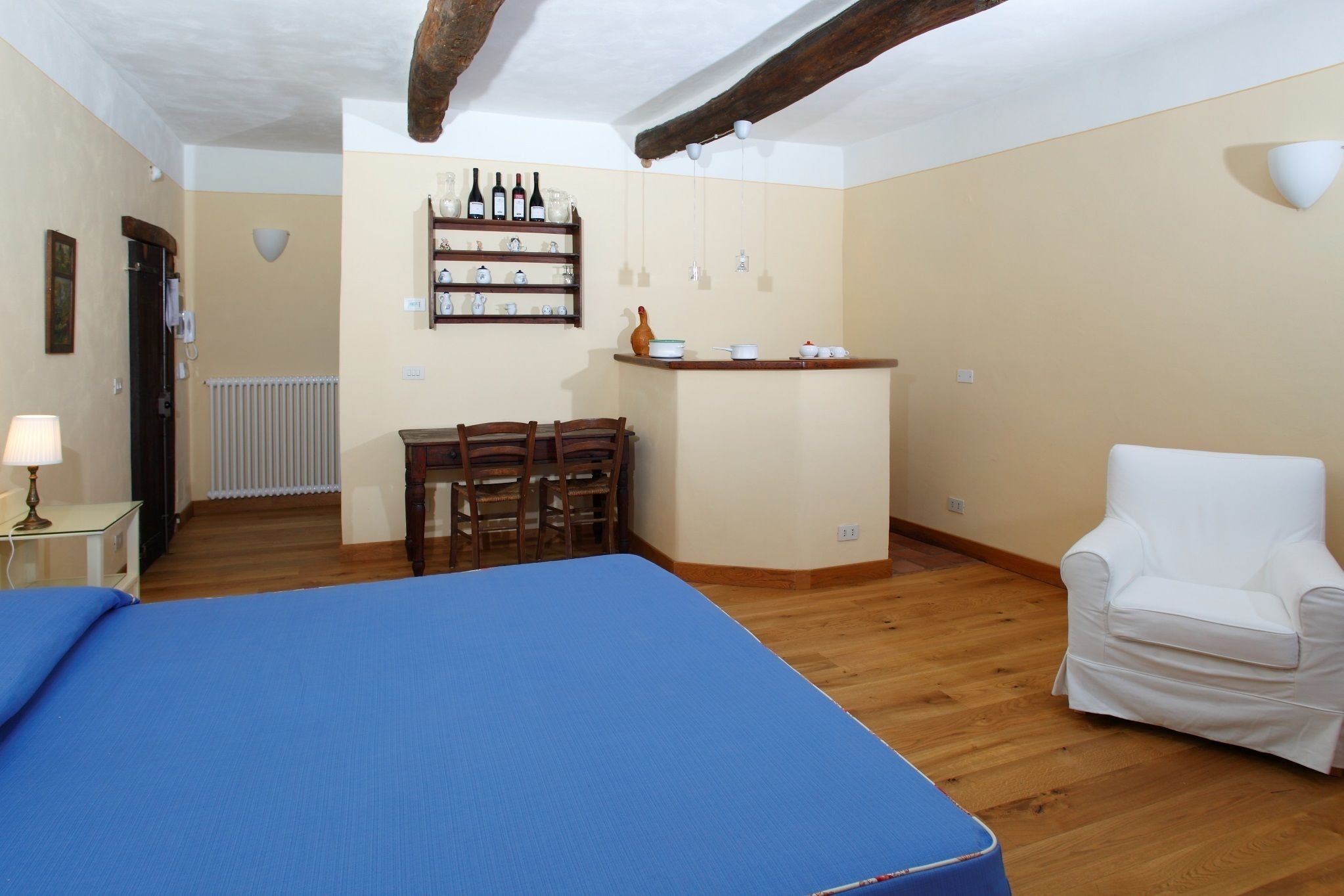 A room with a bed, a chair, a small table, a shelf with bottles, and a radiator.
