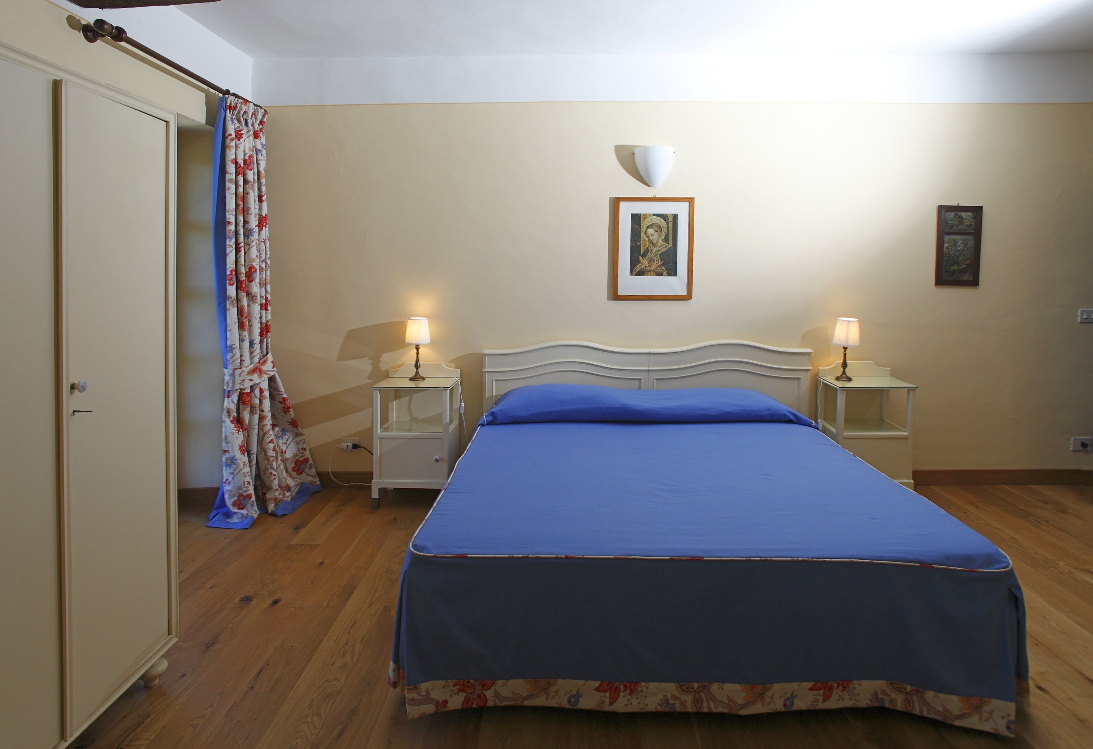 A bedroom with a bed, two bedside tables with lamps, a wardrobe, a curtain, and a framed picture on the wall.