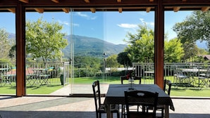 A covered patio with a table and chairs, large glass doors, and a view of a green landscape with trees and mountains.