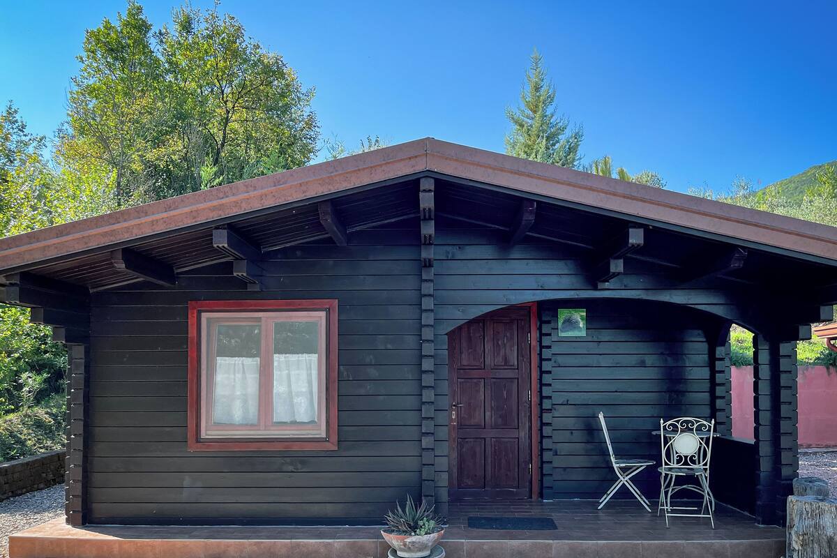 Une petite cabane en bois sombre, avec une fenêtre à cadre rouge et une porte, un petit porche avec une chaise et une table, et un pot de fleurs devant l’entrée.