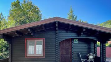 A small, dark wooden cabin with a red-framed window and a door, a small porch with a chair and table, and a potted plant in front.