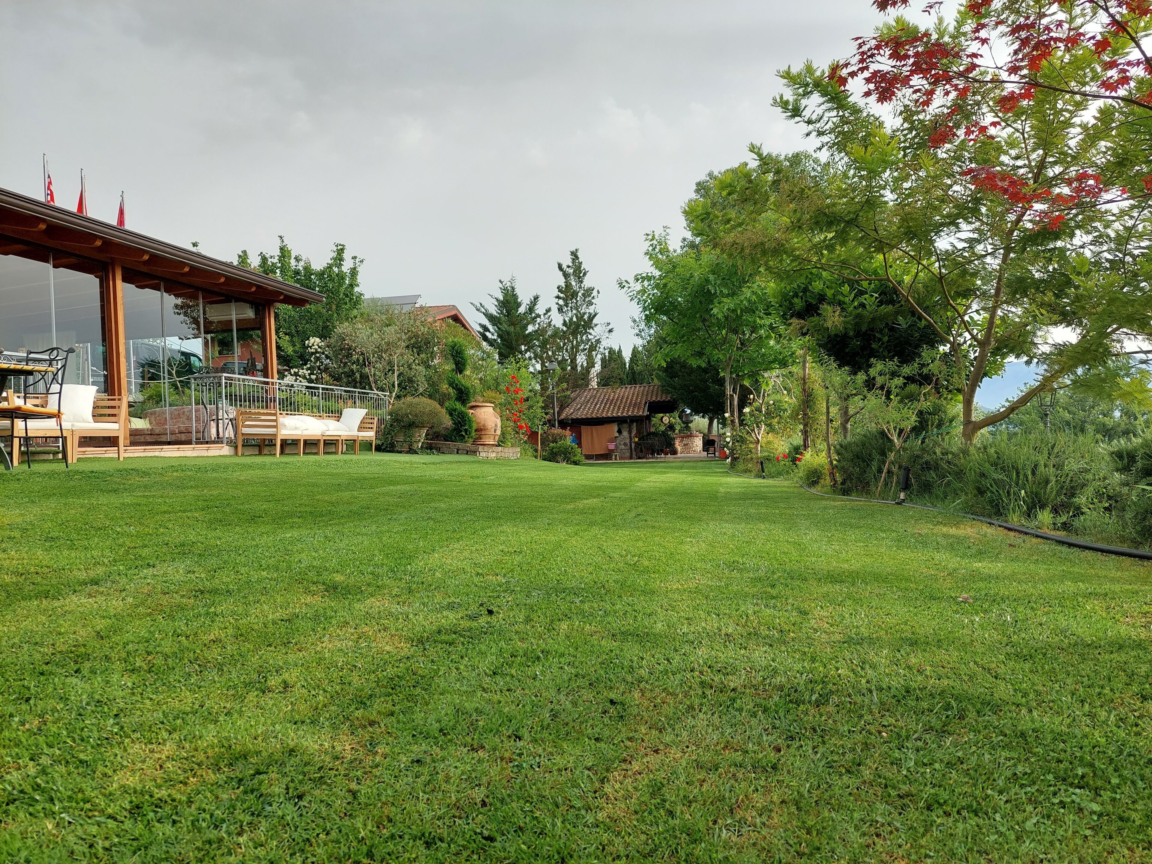 Un césped bien cuidado con mobiliario exterior, una pérgola y una pequeña construcción, presumiblemente una casa de huéspedes o cabaña, rodeado de árboles y vegetación.