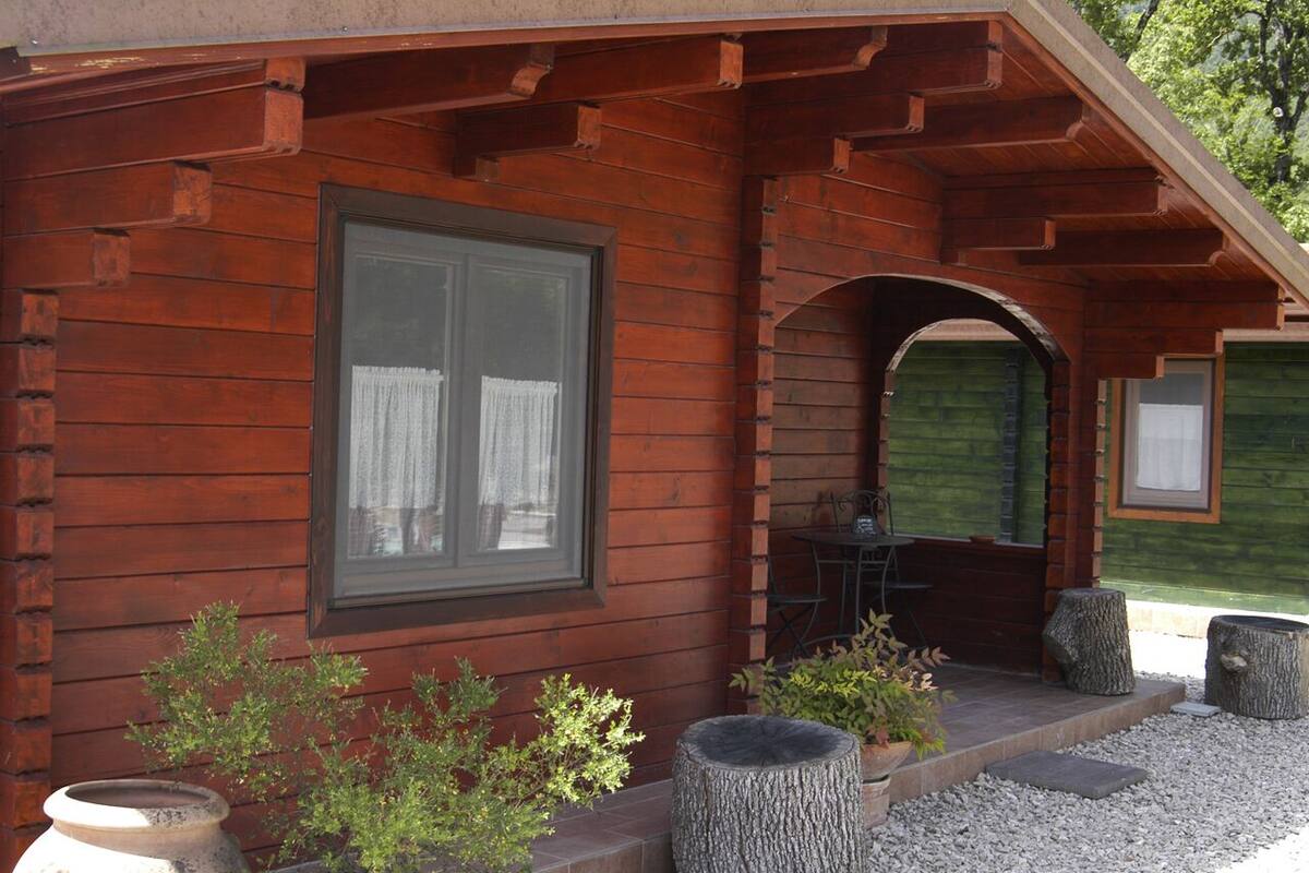 Une cabane en bois dotée d’une grande fenêtre, d’une véranda avec une table et des chaises, et d’un petit jardin agrémenté de plantes en pot et d’une souche d’arbre.