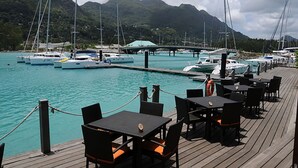 Eine Holzterrasse mit Tischen und Stühlen mit Blick auf einen Jachthafen, wo Boote vor einer Bergkulisse liegen.