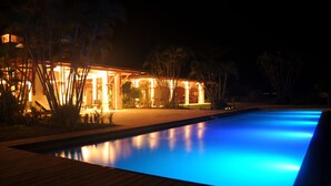 Ein Schwimmbecken mit Holzterrasse, umgeben von Palmen und einem Gebäude mit warmer Beleuchtung.