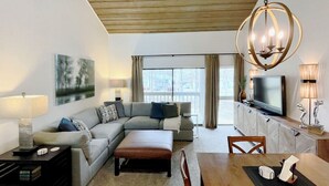 A modern living room with a sectional sofa, a wooden dining table, a TV, and a large window with curtains.