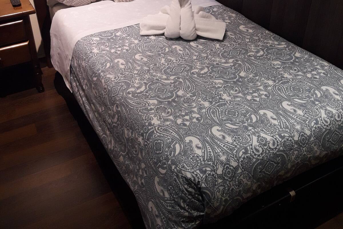 En snyggt bäddad säng med ett mönstrat överkast, kuddar och en hopvikt handduk, placerad på en sängram i trä i ett hotellrum.