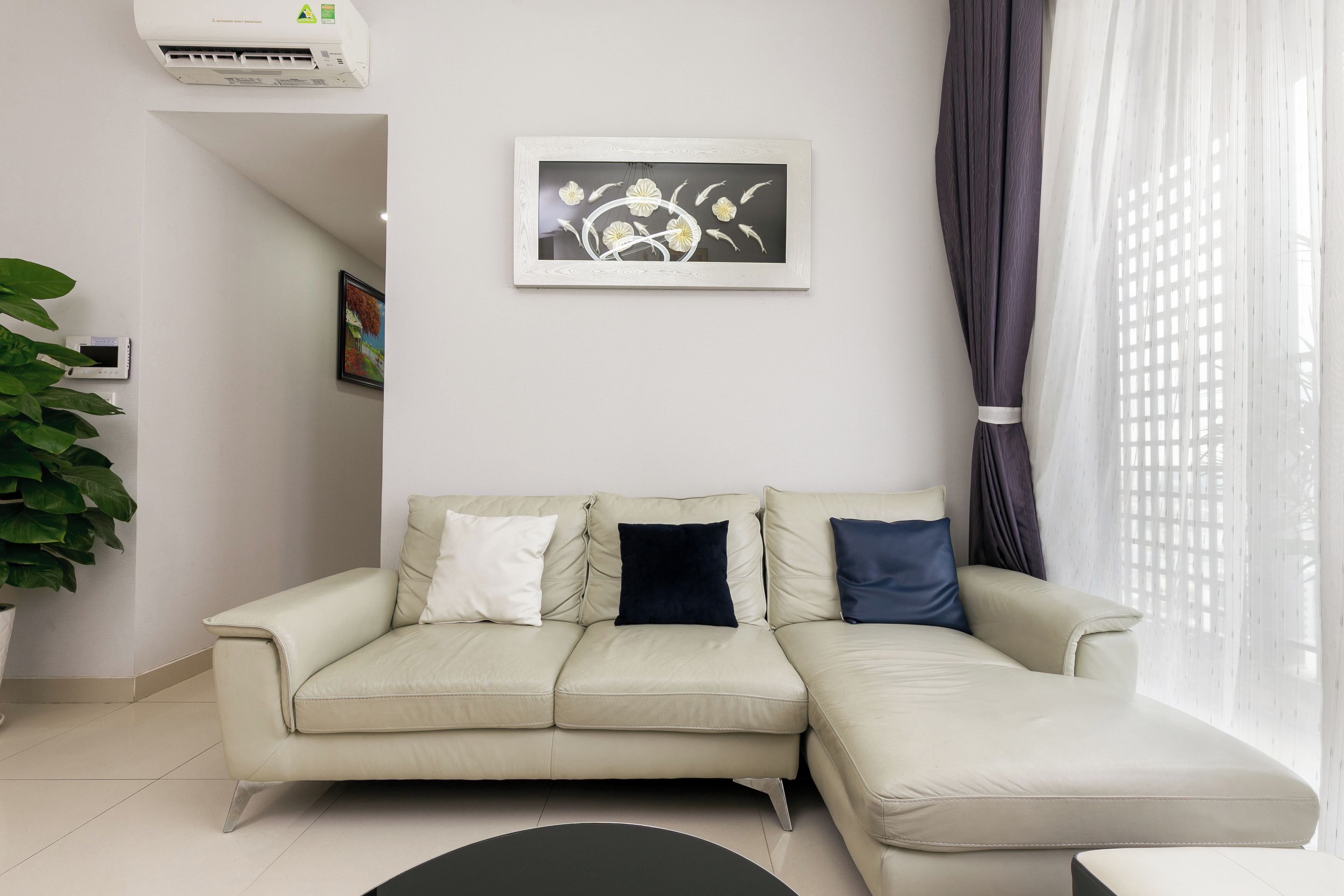 A living room with a beige sofa, a framed artwork, a potted plant, and a window with curtains.