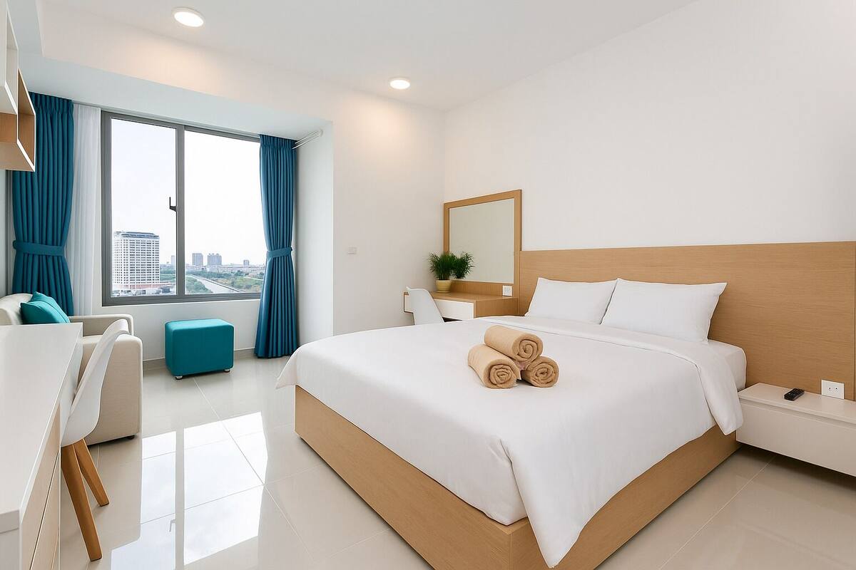 A modern hotel room with a large bed, a desk, a chair, a small table, and a view of the city through large windows.