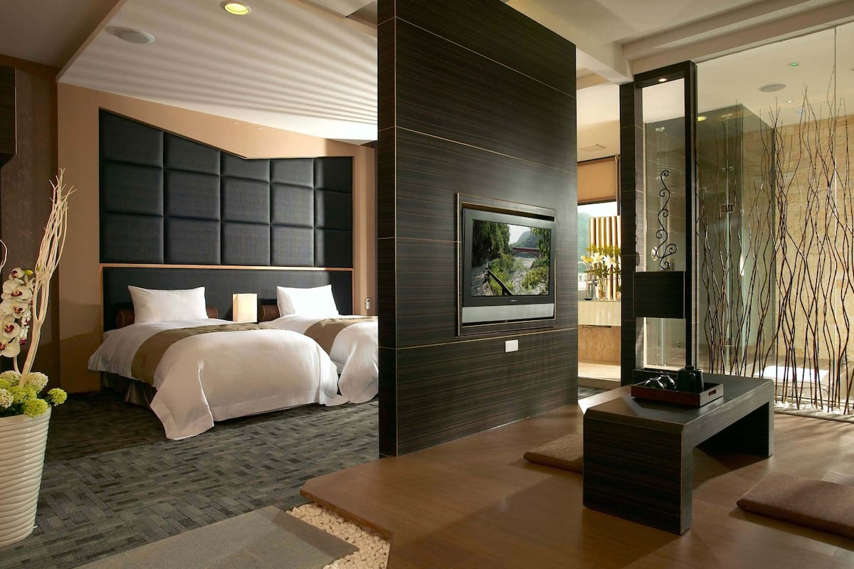 Habitación de hotel moderna con una cama grande, un televisor de pantalla plana montado en una pared oscura, una zona de estar con una mesa de centro y vistas a un puente por la ventana.