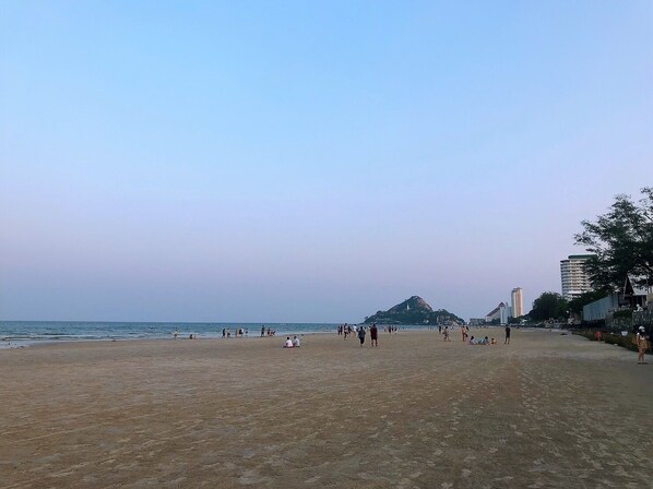 A wide sandy beach with people walking and sitting. There are buildings and a hill in the background.