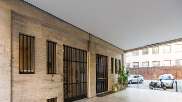 A building with a large open entrance, a covered parking area, and a view of a street with parked cars. - Disciplini (Milan)