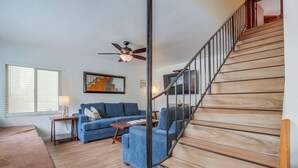 A spacious living room with a wooden staircase, a ceiling fan, and a large window with blinds. - Bring the whole family to this 4 bedroom beach house! (San Diego)