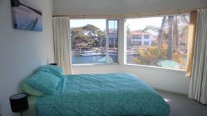 A bedroom with a bed, a nightstand, a lamp, and a window with a view of a body of water and buildings. - Marina's Holiday Retreat (Port Lincoln)