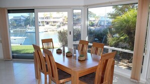 A dining area with a wooden table and chairs, large windows with a view of a pool and greenery. - Marina's Holiday Retreat (Port Lincoln)