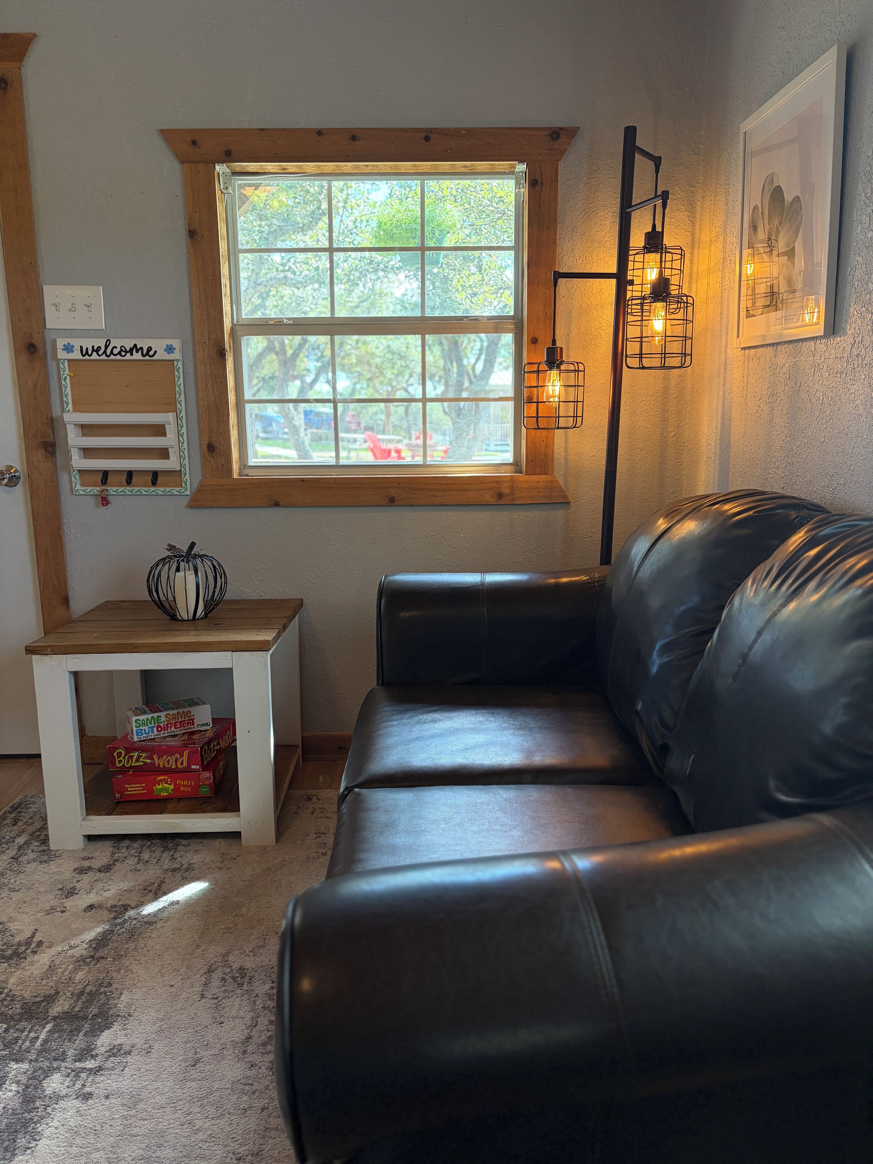 A cozy living room with a leather couch, a wooden side table with board games, a window with a view of trees, and a wall-mounted light fixture.