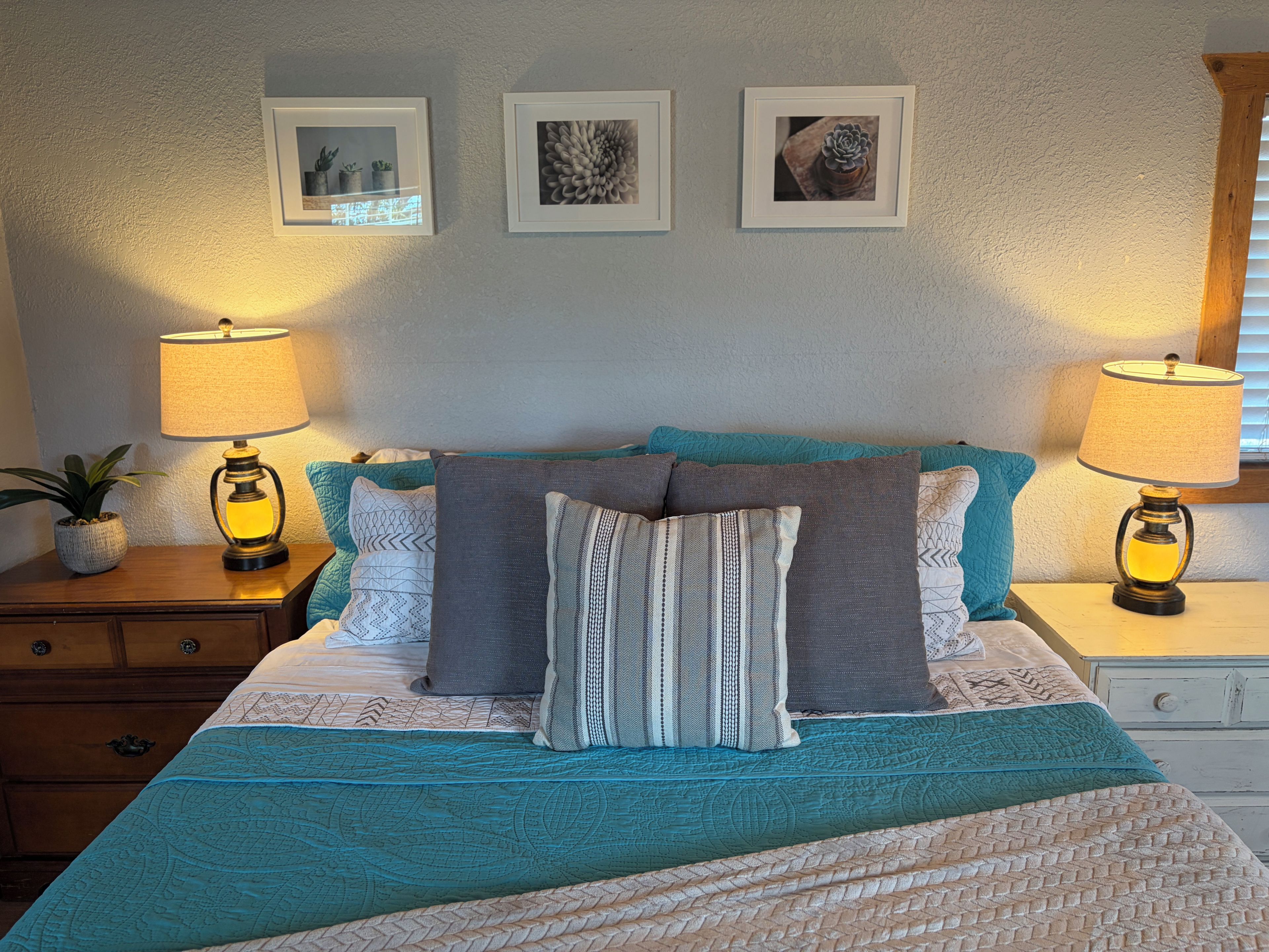 A neatly made bed with a teal and white bedspread, flanked by two bedside lamps, a nightstand with a plant, and a wall with three framed pictures.