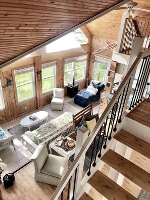 Ein großzügiger Loft-Raum mit hoher Decke, großen Fenstern, einer Holztreppe und einer gemütlichen Sitzecke mit Sofa und Sesseln.