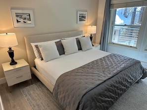 A neatly arranged bedroom with a bed, two bedside tables, a nightstand, a window with blinds, and two framed pictures on the wall.
