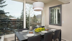 Un espace de repas moderne avec une table dressée pour quatre personnes, de grandes fenêtres offrant une vue sur les arbres et les collines, et un luminaire suspendu au-dessus.
