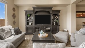 A living room with a large flat-screen TV, dark wood entertainment center, grey sofas, and a coffee table with decorative items.