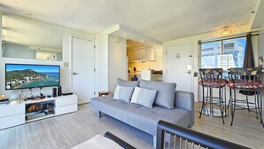 Een moderne woonkamer met een grijze bank, een flatscreen televisie en een eethoek met barkrukken.