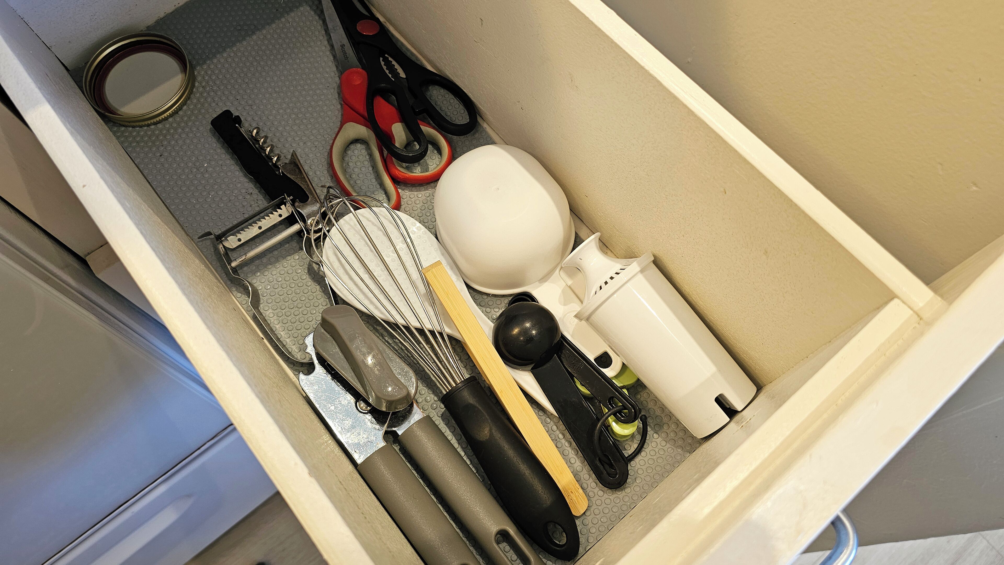 A kitchen drawer with various utensils and appliances including a can opener, scissors, a whisk, a grater, and a white appliance.