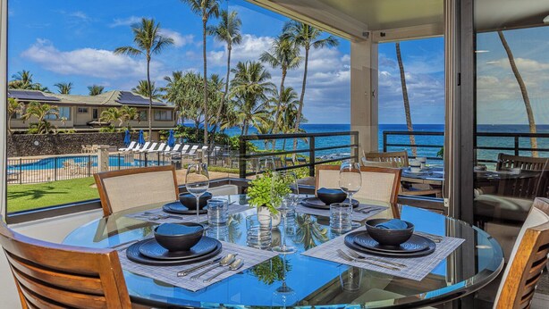 A dining area with a glass table, chairs, and a view of the ocean and palm trees.
