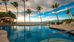 A swimming pool with lounge chairs and a covered seating area, surrounded by palm trees and a view of the ocean.