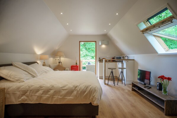 A bedroom with a large bed, a TV, a desk, and a window with a view of greenery.