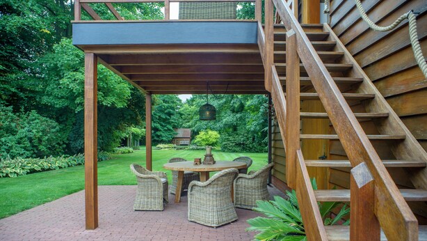 Een overdekt terras met rieten meubilair, een betegelde vloer en een houten trap.