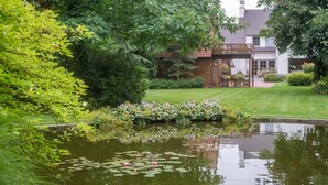 Een rustige tuin met een vijver, weelderige begroeiing en een huis met een houten terras.