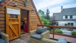 Een houten chalet met een grote open deur, rieten meubels en uitzicht op een tuin en omliggende gebouwen.
