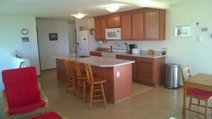 A compact kitchen with wooden cabinets, a microwave, a stove, and a sink. There are three wooden stools around the island, and a trash can is visible.