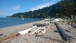 A pebbled beach with boats and logs, surrounded by forested hills.