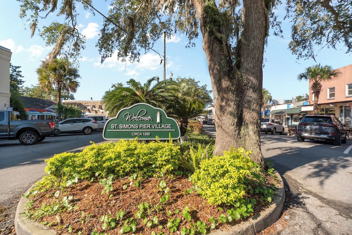 A welcome sign for St. Simons Pier Village with a road and parked cars in the background.