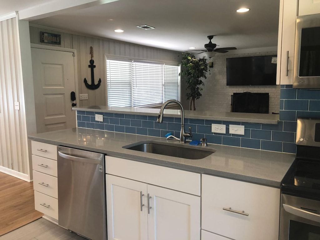 A kitchen with a stainless steel dishwasher, white cabinets, a blue tiled backsplash, and a ceiling fan.
