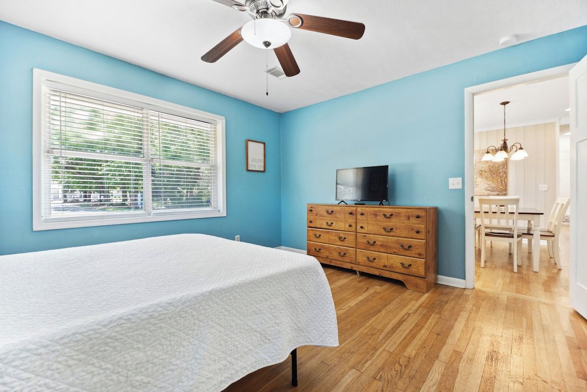 Een slaapkamer met een bed, een dressoir, een plafondventilator en een raam met jaloezieën.