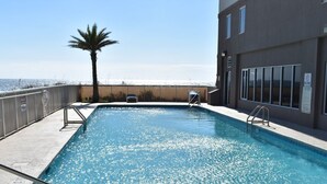 Una piscina rectangular con superficie de agua azul cristalina, rodeada por una solana de concreto. A la izquierda de la piscina hay una palmera, y a la derecha un edificio con ventanas y una puerta.