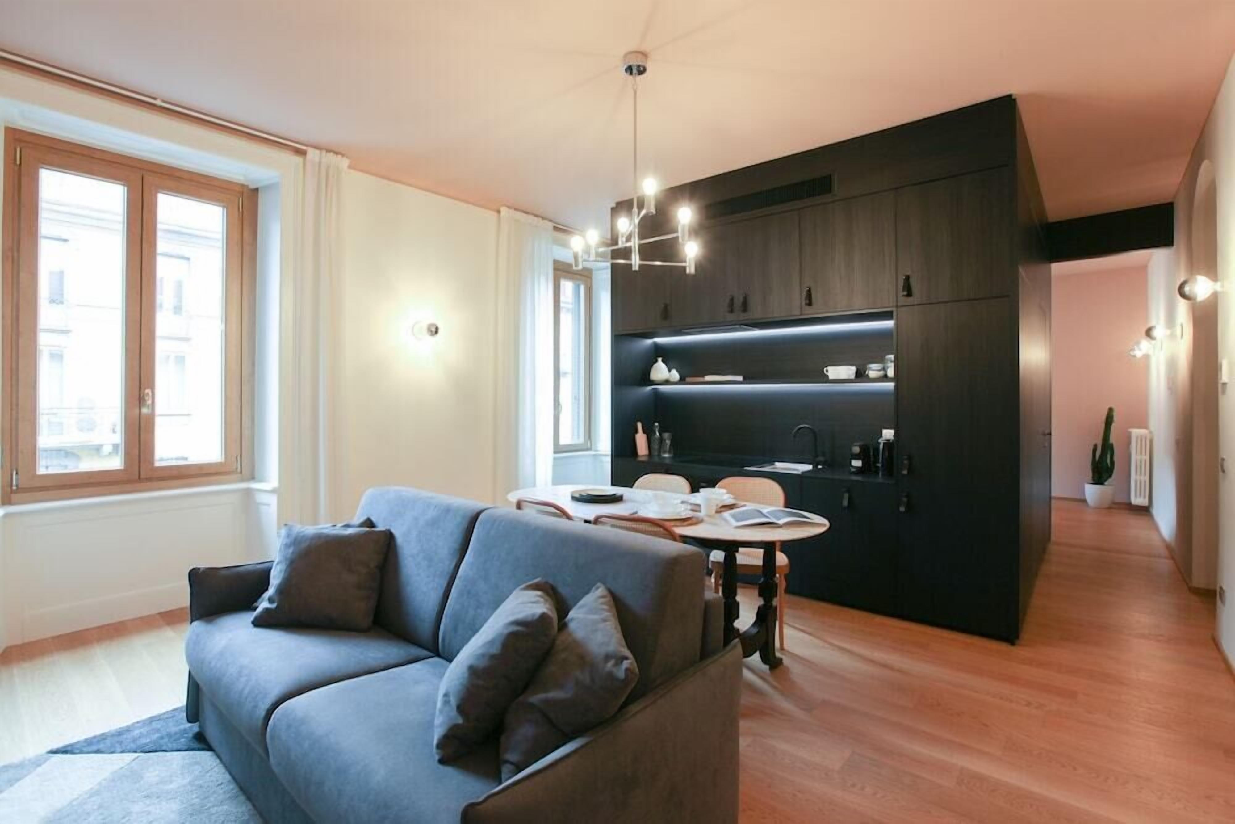 A modern living room with a grey sofa, wooden flooring, and a kitchen area with dark cabinetry and a hanging light fixture.