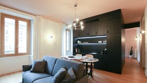 A modern living room with a grey sofa, wooden flooring, and a kitchen area with dark cabinetry and a hanging light fixture. - Vitruvio (Milan)