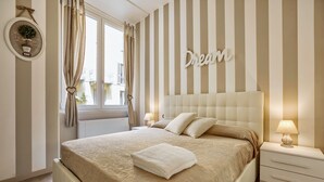 A bedroom with a large bed, striped wallpaper, a bedside table with a lamp, and a window with curtains. - Vitruvio (Milan)