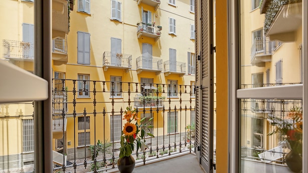 A balcony with a potted sunflower, a metal railing, and a view of yellow buildings with balconies. - Vitruvio (Milan)
