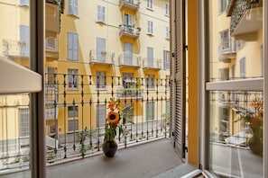 A balcony with a potted sunflower, a metal railing, and a view of yellow buildings with balconies. - Vitruvio (Milan)