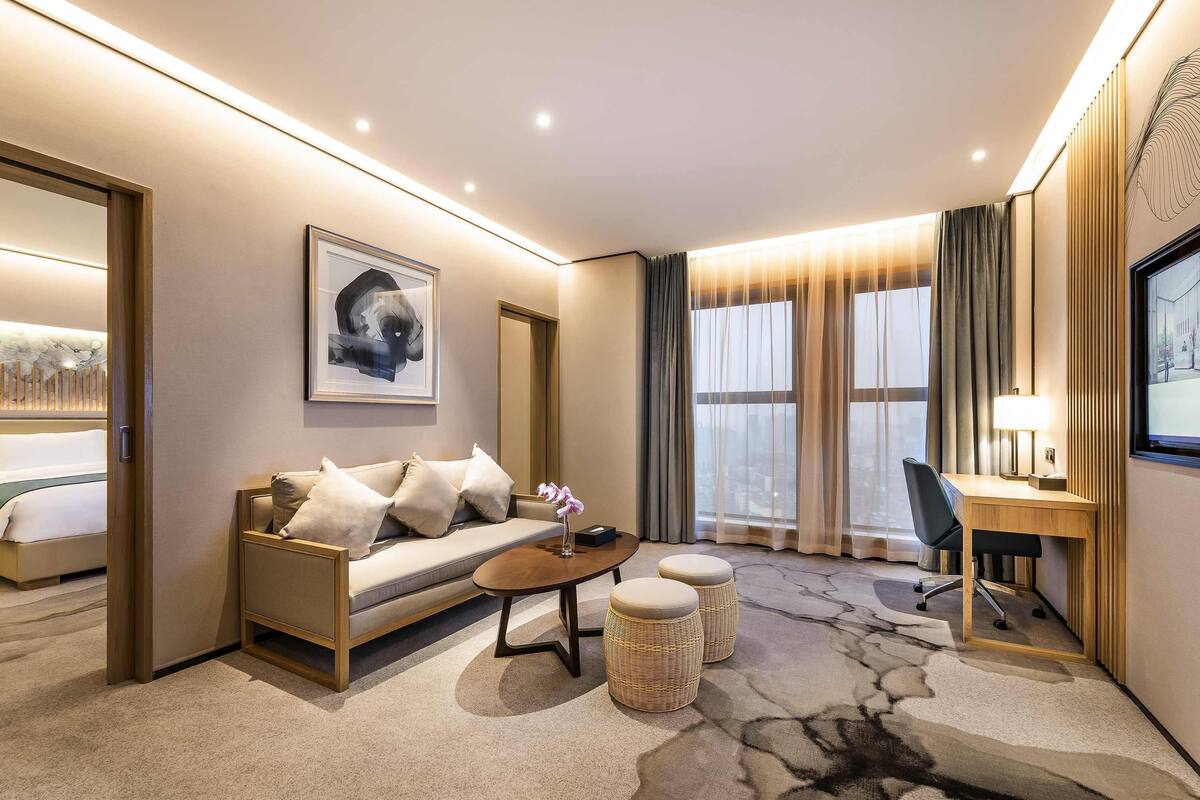 A modern hotel room with a sofa, a desk, a chair, a coffee table, and a large window with curtains.